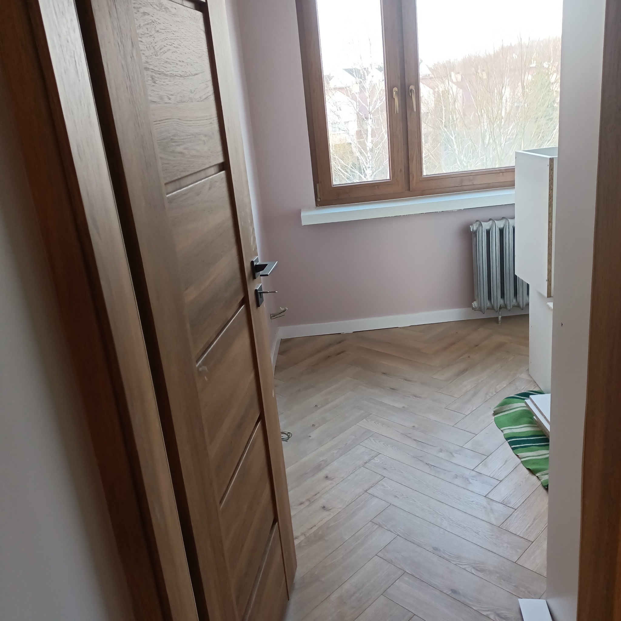 Jasne, drewniane panele ułożone w jodełkę, widoczne drzwi z ciemnego drewna, fragment okna z grzejnikiem i fragment mebla do montażu, różowe ściany.