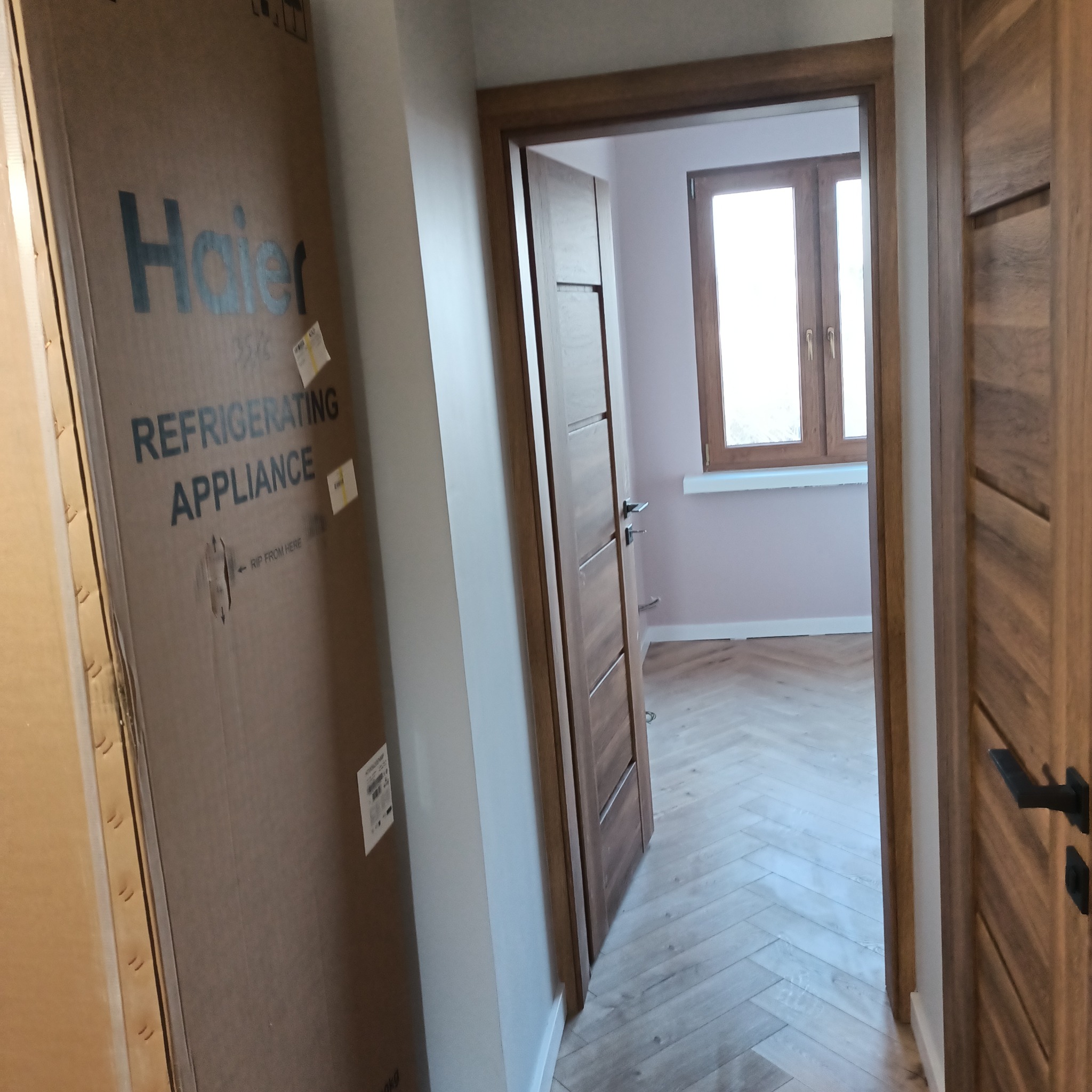Nowe panele ułożone we wzór jodełki francuskiej, widoczne przez otwarte drzwi z drewnianą ościeżnicą, obok stoi karton z nowym sprzętem AGD.
