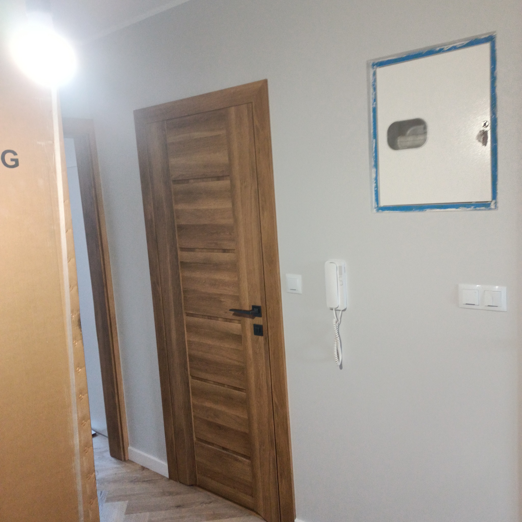 Nowo zamontowane drewniane drzwi wewnętrzne z czarną klamką, widoczne na tle szarej ściany z domofonem i skrzynką elektryczną w niebieskiej ramce, obok karton z częściowo widocznym napisem.