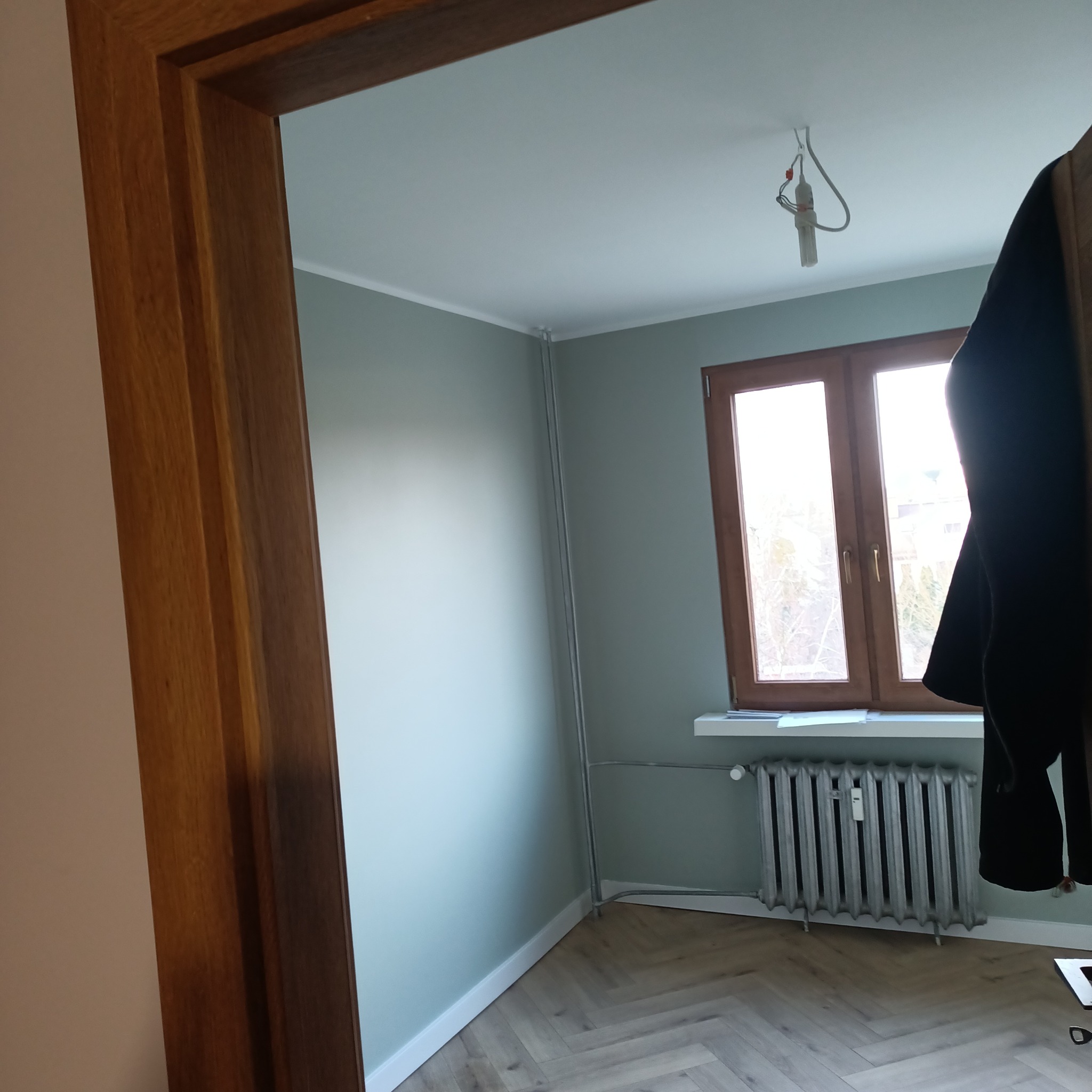 Wnętrze pokoju po remoncie: świeżo pomalowane ściany w odcieniach zieleni, podłoga wyłożona panelami w jodełkę, drewniane okno i grzejnik żeliwny.