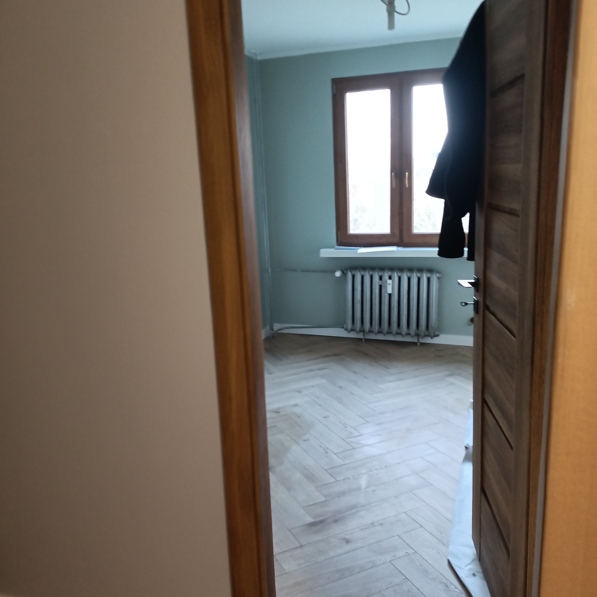 Widok na pokój z ułożonymi jasnymi panelami podłogowymi w jodełkę, przez uchylone drewniane drzwi, z widocznym grzejnikiem i oknem w tle.