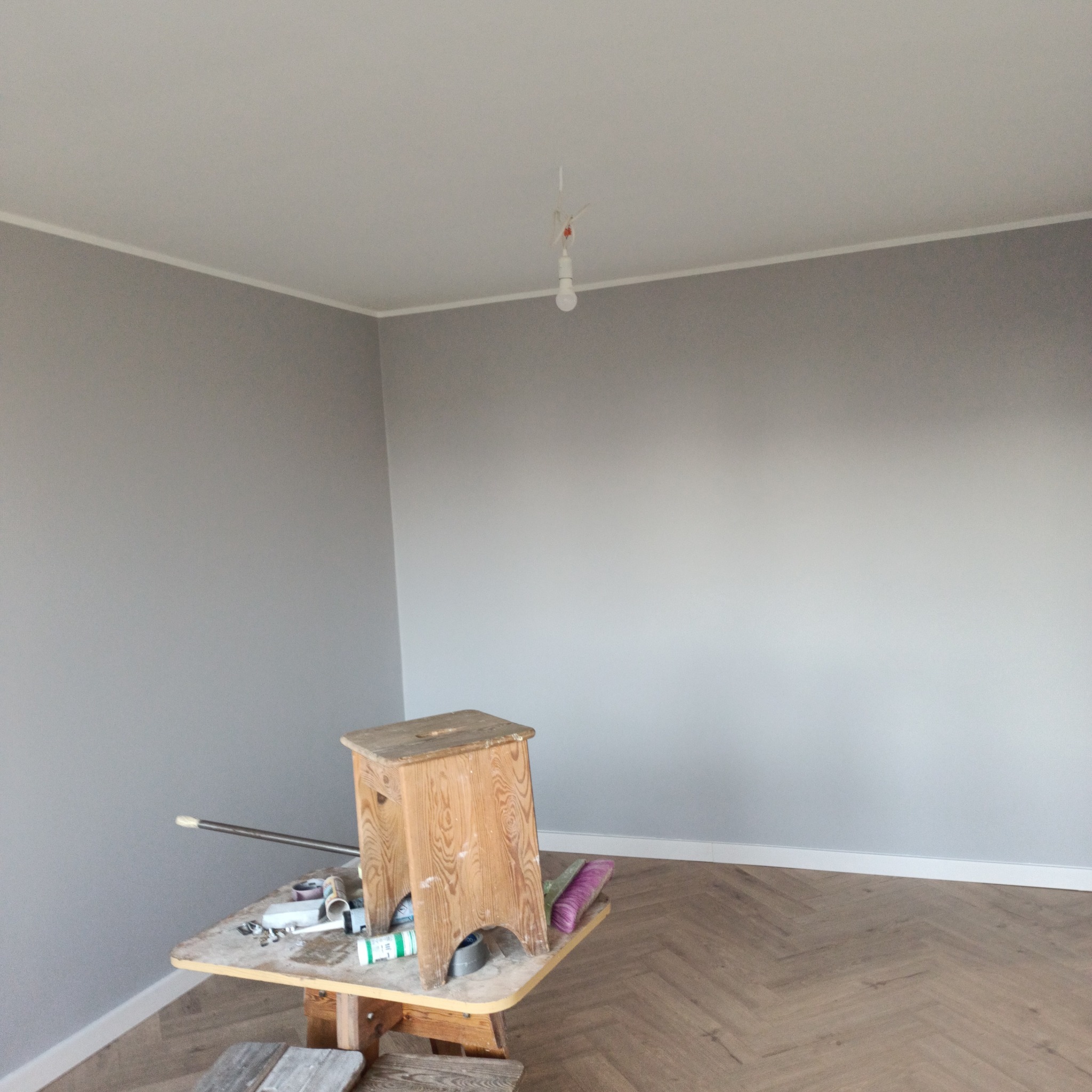 Pokój po świeżym malowaniu na szaro, z drewnianą podłogą ułożoną w jodełkę, widoczny stolik z narzędziami malarskimi i drabiną, surowa żarówka zwisająca z sufitu.