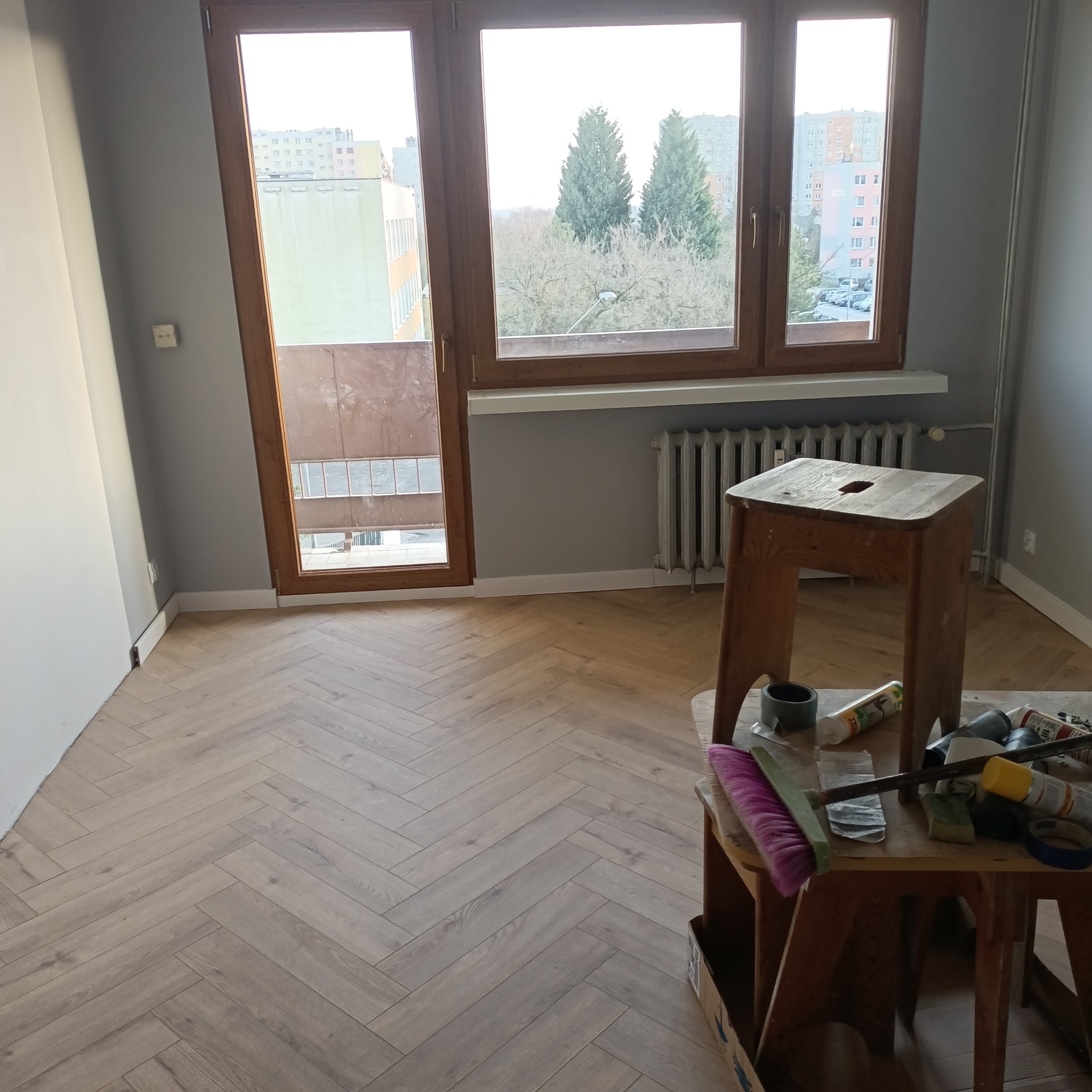 Jasne panele ułożone w jodełkę francuską w pokoju z szarymi ścianami, widok z okna na budynki i drzewa, na pierwszym planie taboret z narzędziami i materiałami malarskimi.