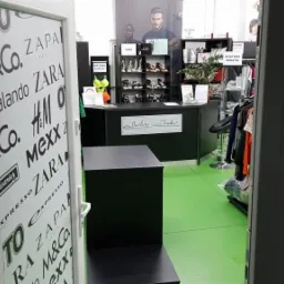 Wnętrze sklepu odzieżowego z zieloną podłogą, widoczne logo marek na szklanych drzwiach wejściowych, lada z napisem 'Outlety Trade' i półki z obuwiem oraz manekiny z ubraniami w tle.