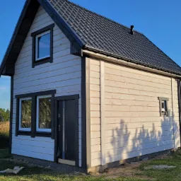 Niewielki, jasny budynek z drewnianą elewacją i ciemnym dachem z blachodachówki, widoczne okna z białymi ramami i ciemne obramowania, teren wokół w trakcie zagospodarowywania.