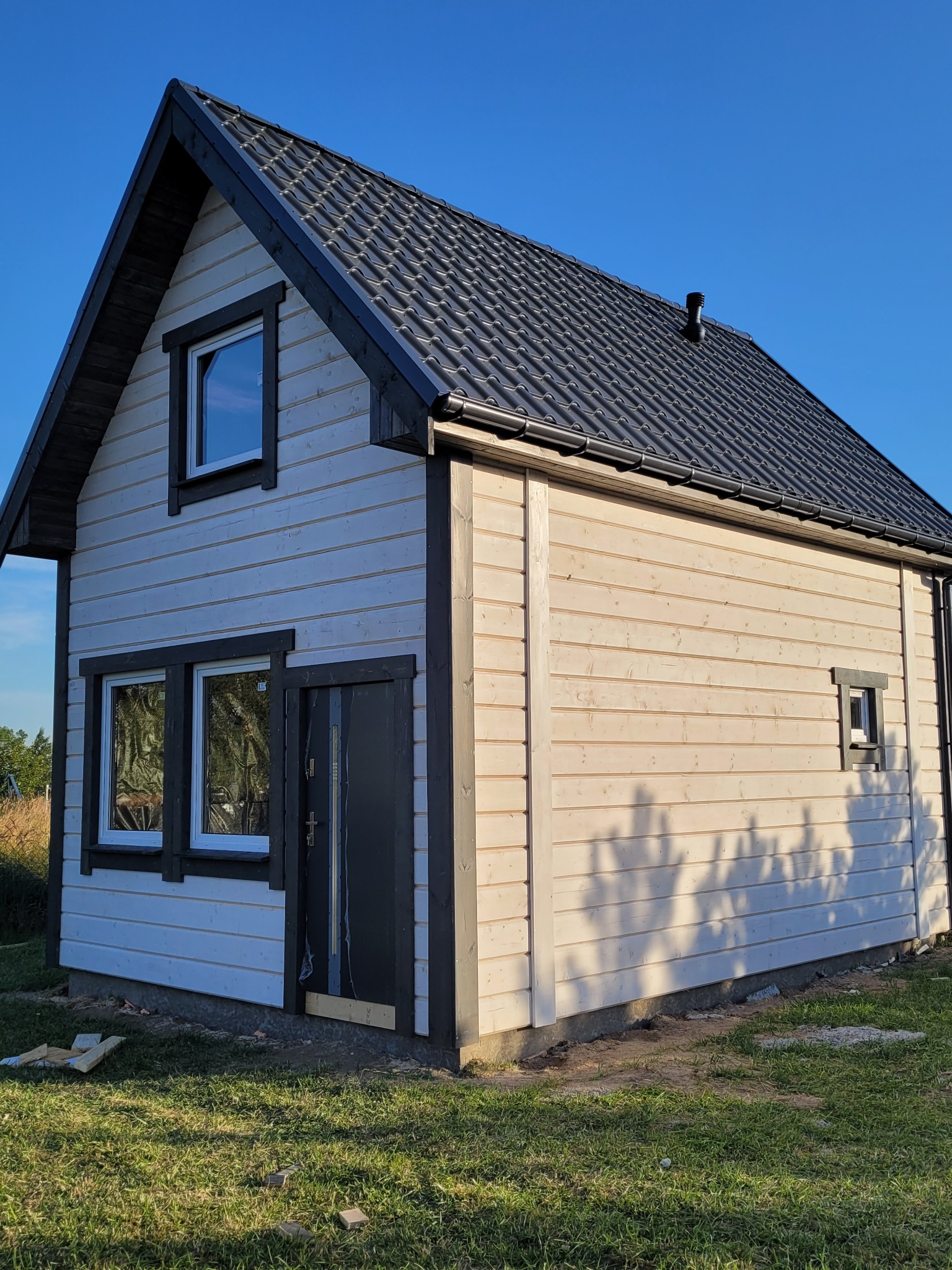 Niewielki, jasny budynek z drewnianą elewacją i ciemnym dachem z blachodachówki, widoczne okna z białymi ramami i ciemne obramowania, teren wokół w trakcie zagospodarowywania.