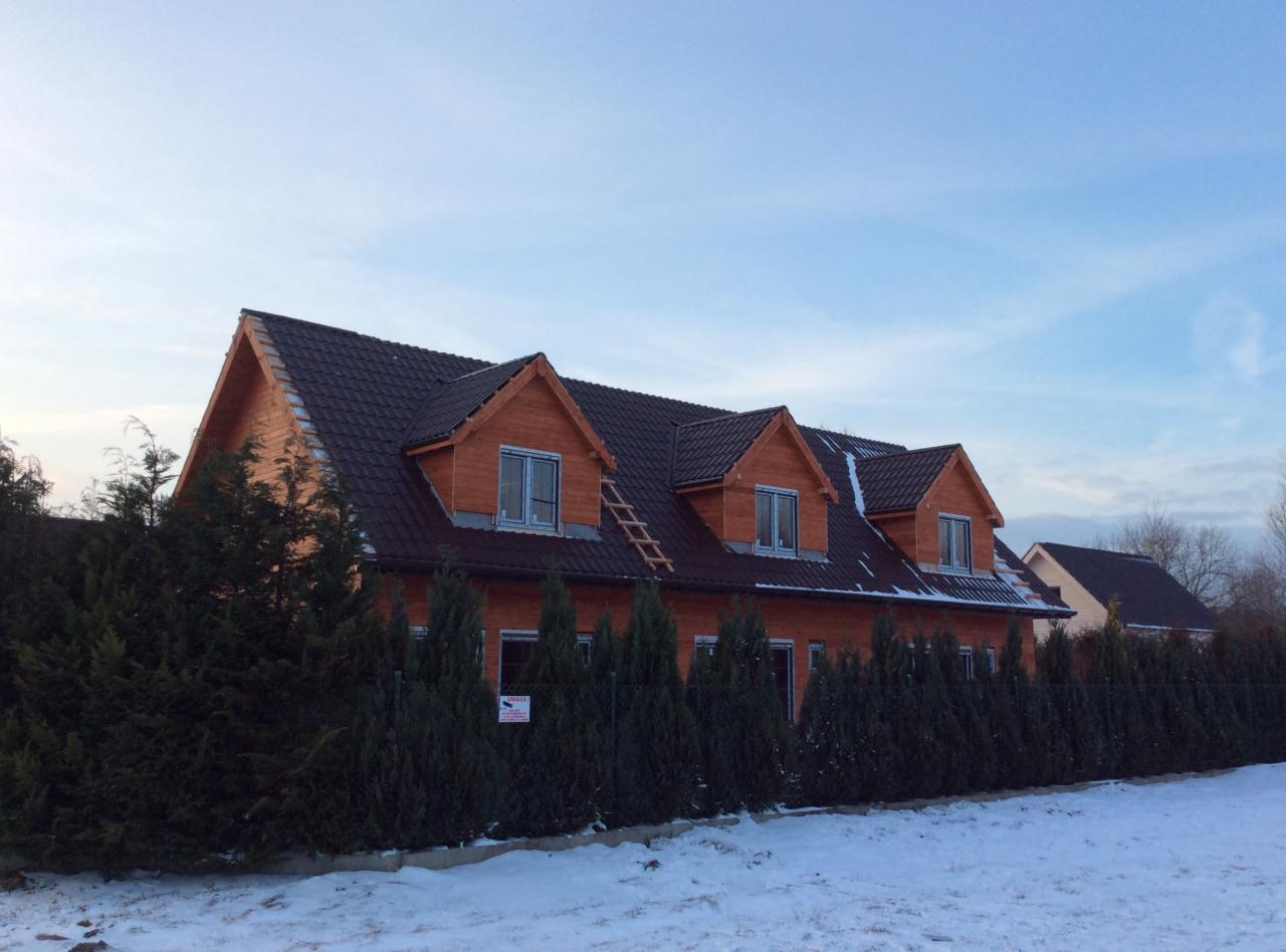 Gotowy dom z drewna, z brązowym dachem i trzema oknami dachowymi, drabina oparta o dach, otoczony tujami i ogrodzeniem z siatki, zimowy krajobraz.