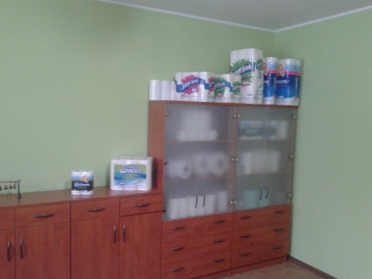 Zestaw papieru toaletowego i ręczników papierowych różnych marek, ułożony na szafkach w pomieszczeniu o zielonych ścianach. Widoczne opakowania White Swan i Colore.