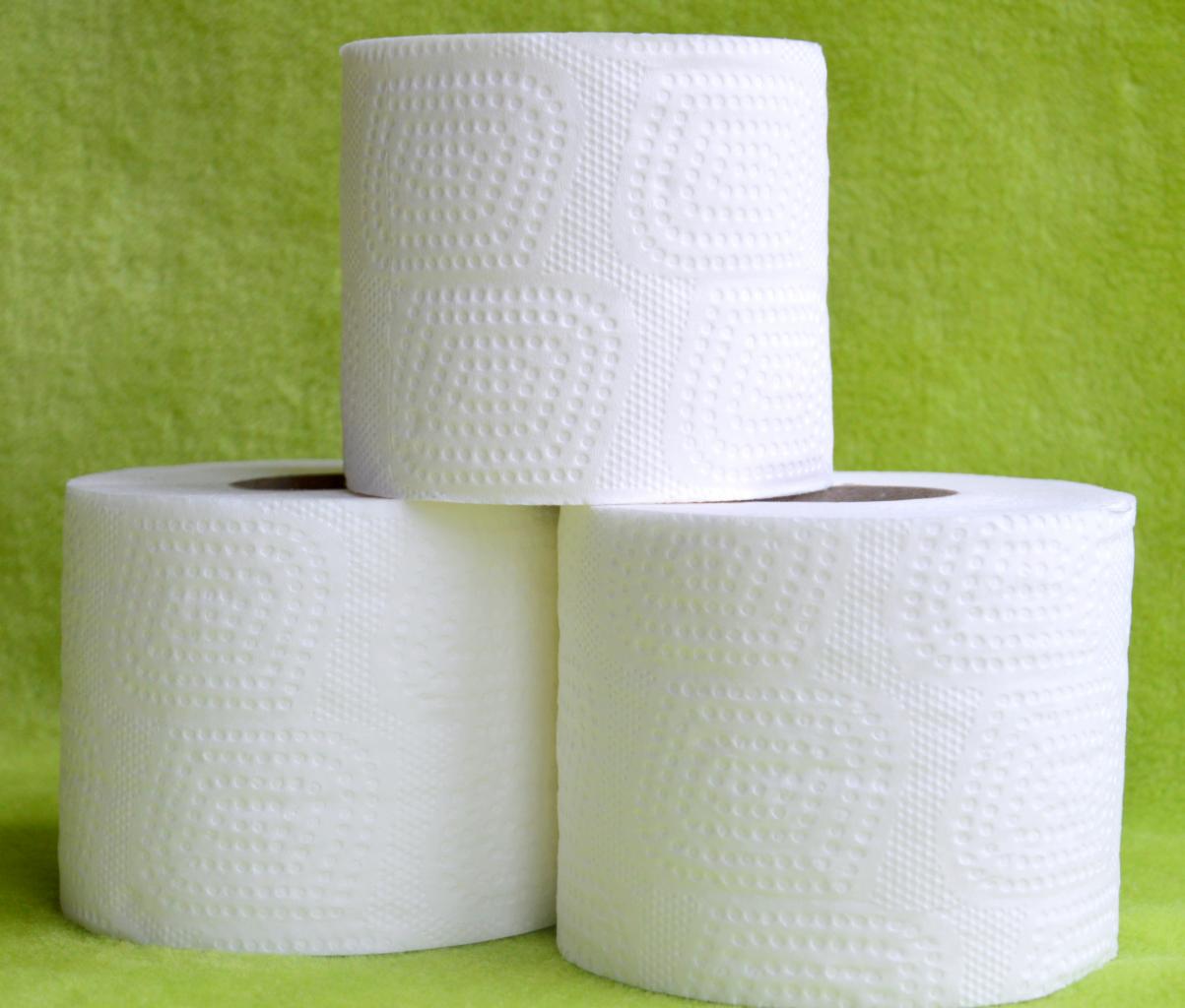 Trzy rolki białego, wzorzystego papieru toaletowego ułożone jedna na drugiej na tle zielonej, teksturowanej powierzchni.