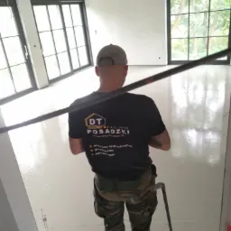 Pracownik w kamuflażowych spodniach wyrównuje świeżo wylaną, jasną posadzkę w przestronnym wnętrzu z dużymi oknami, widoczny sprzęt do rozprowadzania masy i logo firmy na koszulce.