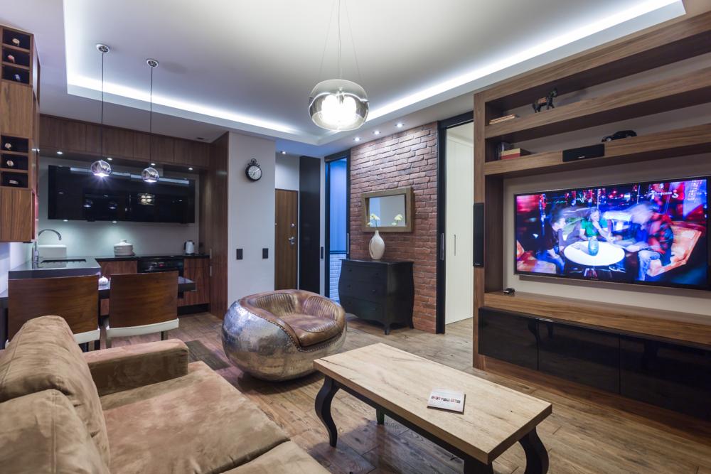 Nowoczesny salon z ceglaną ścianą, designerskim fotelem, drewnianą podłogą i sufitem podwieszanym z taśmą LED, oświetlony wiszącą lampą kulą i punktowymi lampami nad aneksem kuchennym.