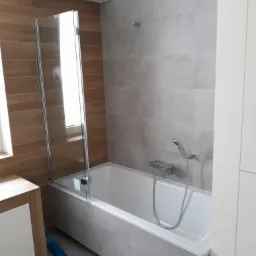 Nowoczesna łazienka z wanną obudowaną szarymi płytkami, szklanym parawanem nawannowym i brązowymi płytkami imitującymi drewno na ścianie. Widoczna bateria wannowa z słuchawką prysznicową i fragment...