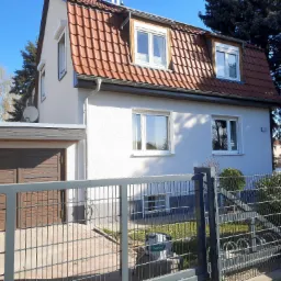 Jasny, otynkowany dom jednorodzinny z czerwoną dachówką, widziany zza metalowego ogrodzenia w słoneczny dzień.