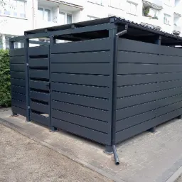 Nowoczesna, szara obudowa śmietnikowa z poziomych paneli, z metalowym dachem i rynną, na kostce brukowej, z zielonym żywopłotem i budynkiem w tle.