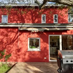 Czerwony dom z drewnianym tarasem, na którym stoi grill. Na elewacji widoczne cienie drzew, okna z białymi ramami i dekoracyjne lampki. W tle widoczny fragment wnętrza domu.