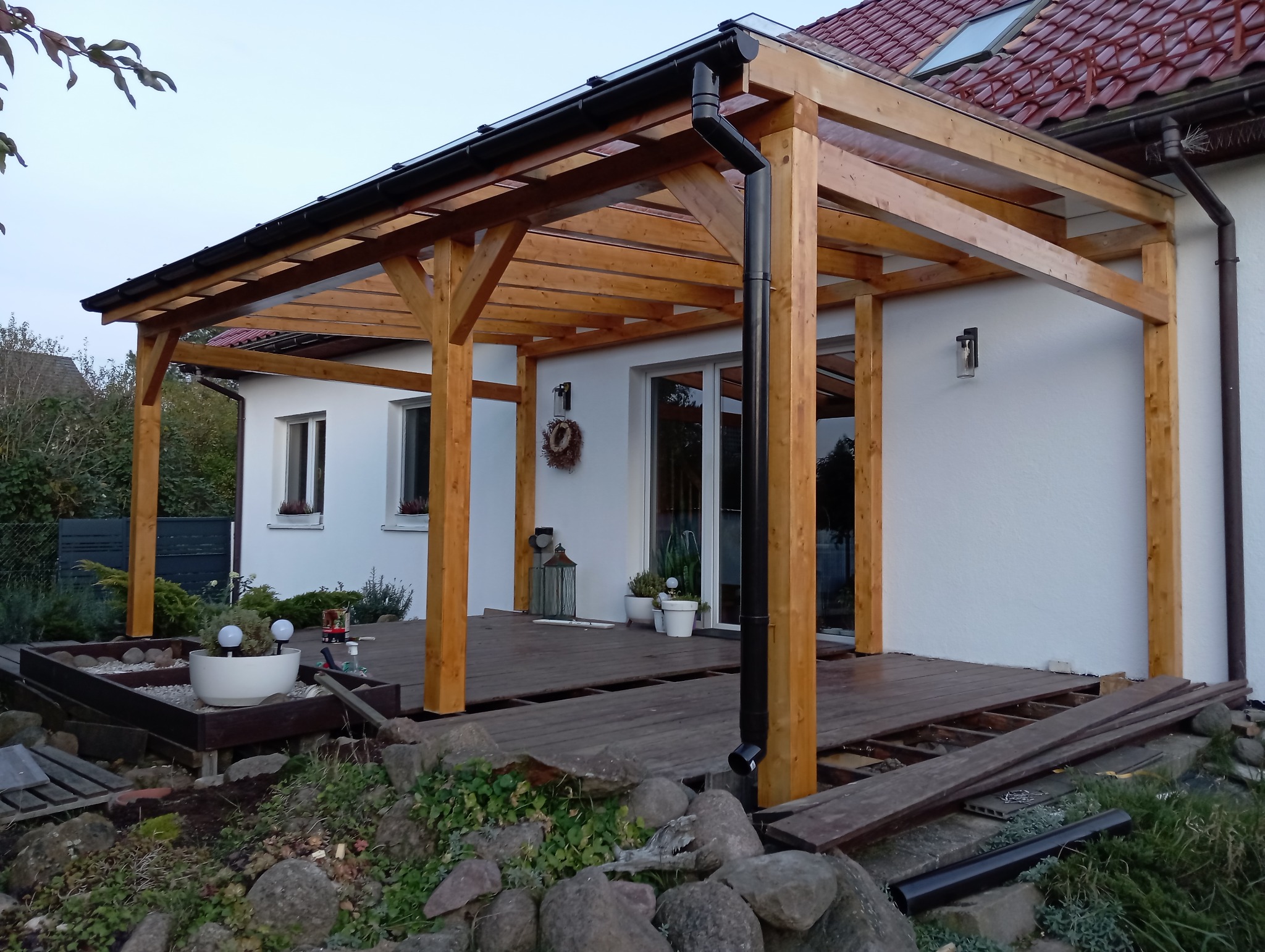 Drewniana konstrukcja zadaszenia tarasu w trakcie budowy, widoczne niekompletne deski podłogowe i elementy dekoracyjne w ogrodzie.