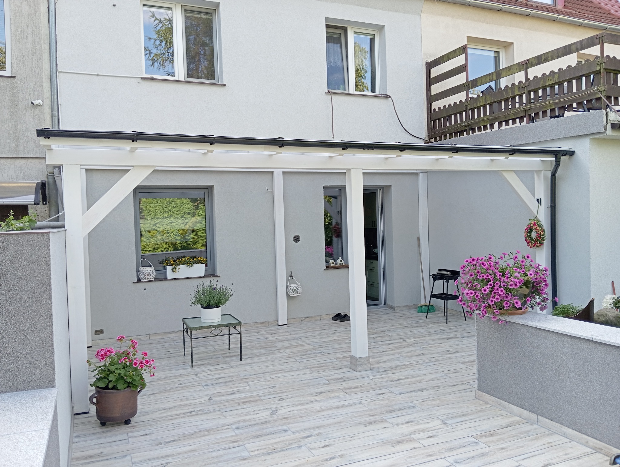 Biała pergola z czarnym zadaszeniem przy szarym domu z oknami, dekorowana kwiatami w donicach i wiankiem, na tarasie z płytek imitujących drewno.