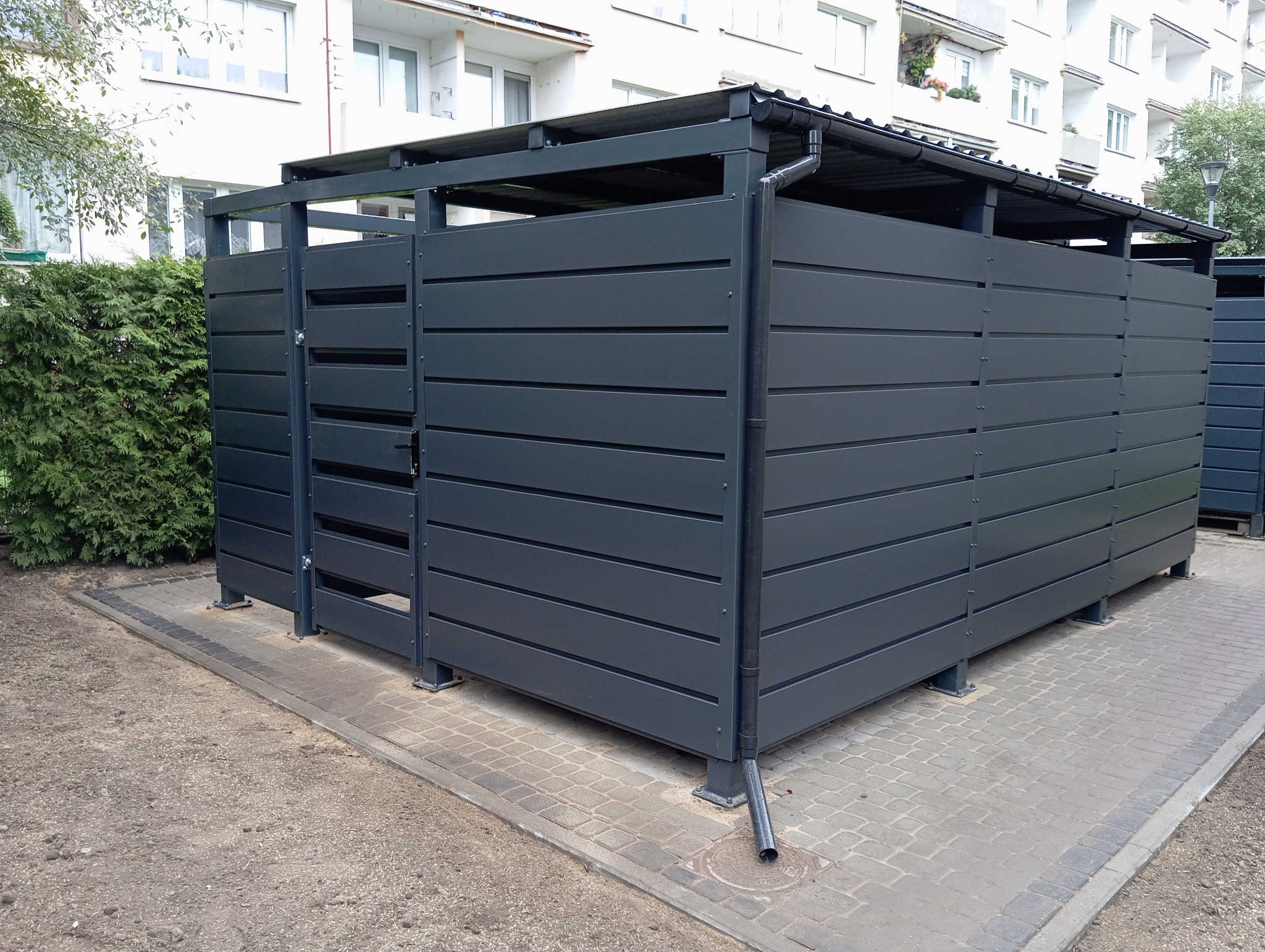 Nowoczesna, szara obudowa śmietnikowa z poziomych paneli, z metalowym dachem i rynną, na kostce brukowej, z zielonym żywopłotem i budynkiem w tle.