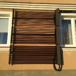 Kolektor słoneczny rurowy zamontowany na elewacji budynku, częściowo zasłaniający okno, z widocznymi rurami przyłączeniowymi. Jasne tło elewacji.