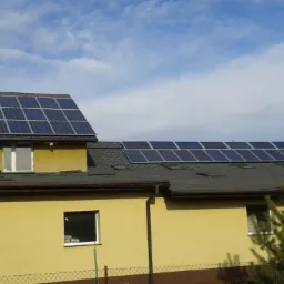 Żółty budynek z dwoma zestawami paneli słonecznych na dachu, widoczne okna i fragment ogrodu. Instalacja fotowoltaiczna na dachu budynku mieszkalnego.