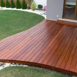 Zakrzywiony taras z egzotycznego drewna, otoczony białym żwirem i zielonym trawnikiem. Widok z g&oacute;ry na harmonijne połączenie natury i architektury.