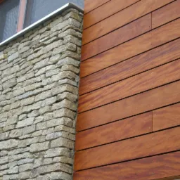 Elewacja budynku z drewnianą okładziną i kamienną ścianą. Widoczny fragment okna i metalowe wykończenie. Detal łączenia materiałów.