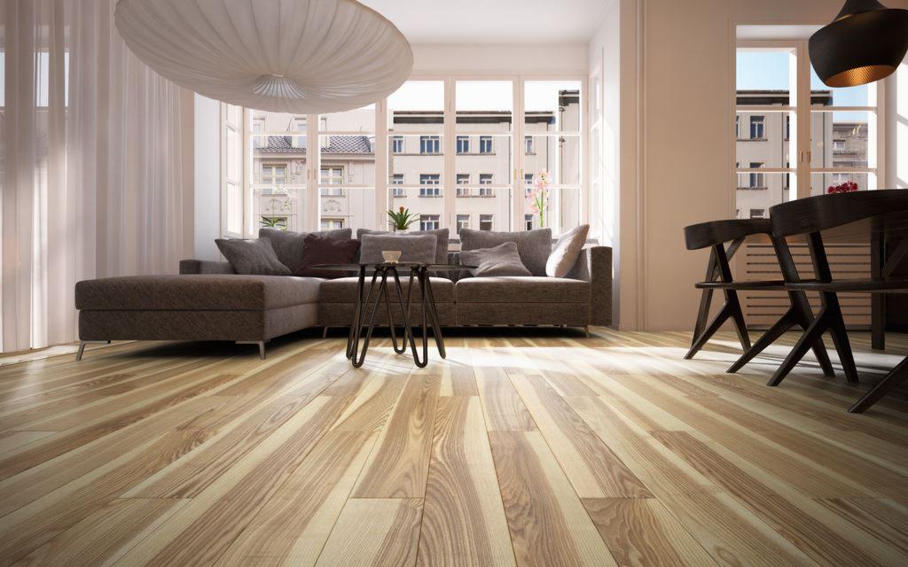 Jasna, drewniana podłoga w salonie z szarym narożnikiem i widokiem na budynek przez okno. Parkiet ułożony w deski, widoczne słoje drewna. Nowoczesne krzesła w tle.