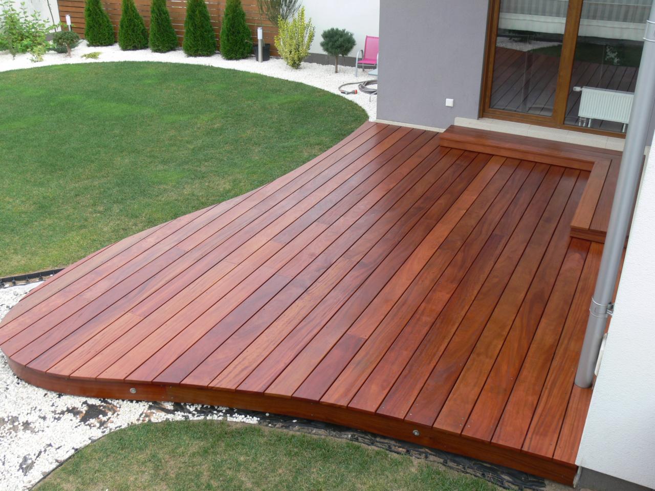 Zakrzywiony taras z drewna egzotycznego, otoczony białym żwirem i zielonym trawnikiem. Widok z góry, podkreślający unikalny kształt i wysoką jakość wykonania.