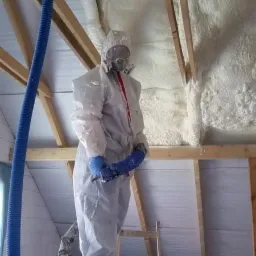 Pracownik w kombinezonie ochronnym aplikuje izolację piankową na poddaszu. Widoczna niebieska rura doprowadzająca materiał i drewniana konstrukcja dachu.