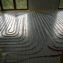 Białe rurki ogrzewania podłogowego ułożone w pętle na siatce montażowej, widoczne czerwone końcówki rur, duże okna w tle.