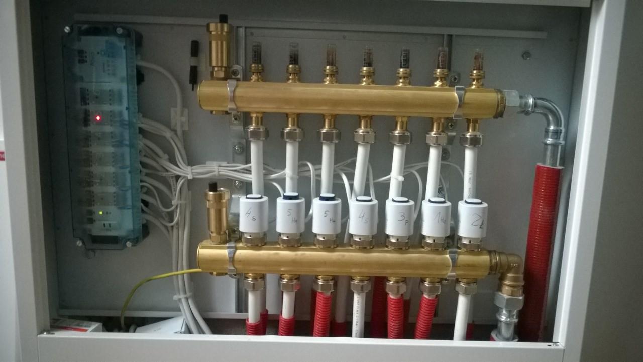 Rozdzielacz ogrzewania podłogowego w szafce z widocznymi rurami, zaworami regulacyjnymi z opisem stref i sterownikiem elektronicznym.