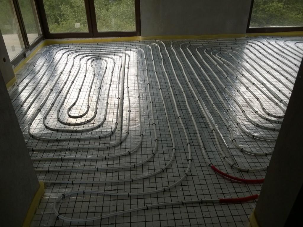 Białe rurki ogrzewania podłogowego ułożone w pętle na siatce montażowej, widoczne czerwone końcówki rur, duże okna w tle.