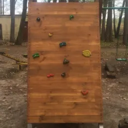 Drewniana ścianka wspinaczkowa dla dzieci z kolorowymi uchwytami, ustawiona na zewnątrz w otoczeniu drzew i placu zabaw.
