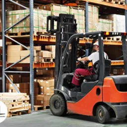 Operator w czerwonych spodniach prowadzi pomarańczowy wózek widłowy w magazynie z regałami wypełnionymi kartonami.