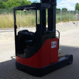 Wózek widłowy wysokiego składowania Linde R16, czerwono-czarny, zaparkowany na kostce brukowej przed ogrodzeniem z siatki i zielenią.