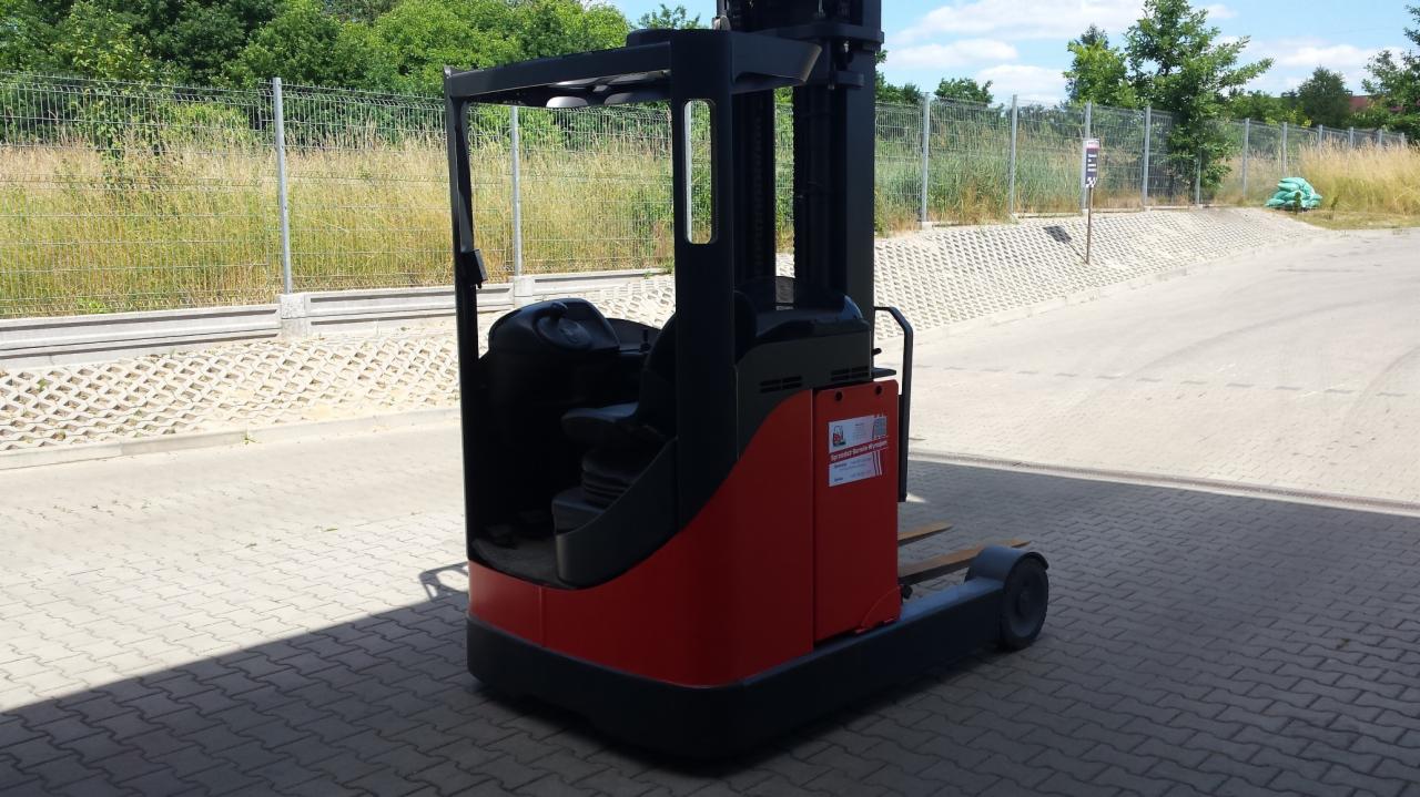 Wózek widłowy wysokiego składowania Linde R16, czerwono-czarny, zaparkowany na kostce brukowej przed ogrodzeniem z siatki i zielenią.