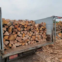 Kłody drewna kominkowego załadowane do skrzyni samochodu ciężarowego z widocznym żurawiem do załadunku na tle składowanych pni drzew.