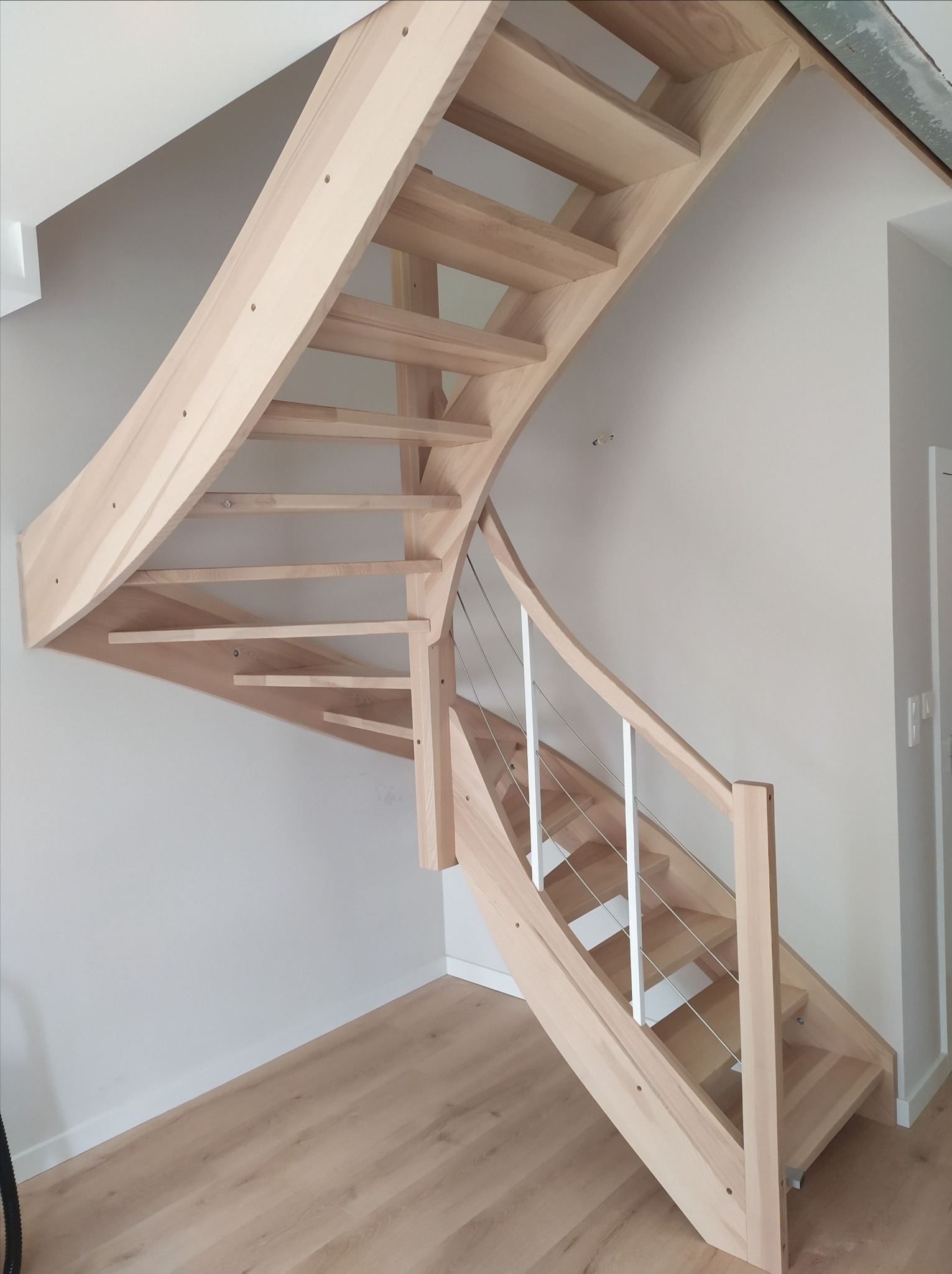 Jasne, gięte schody z drewna z białymi balustradami i stalowymi linkami, widok z dołu na tle jasnych ścian i drewnianej podłogi.