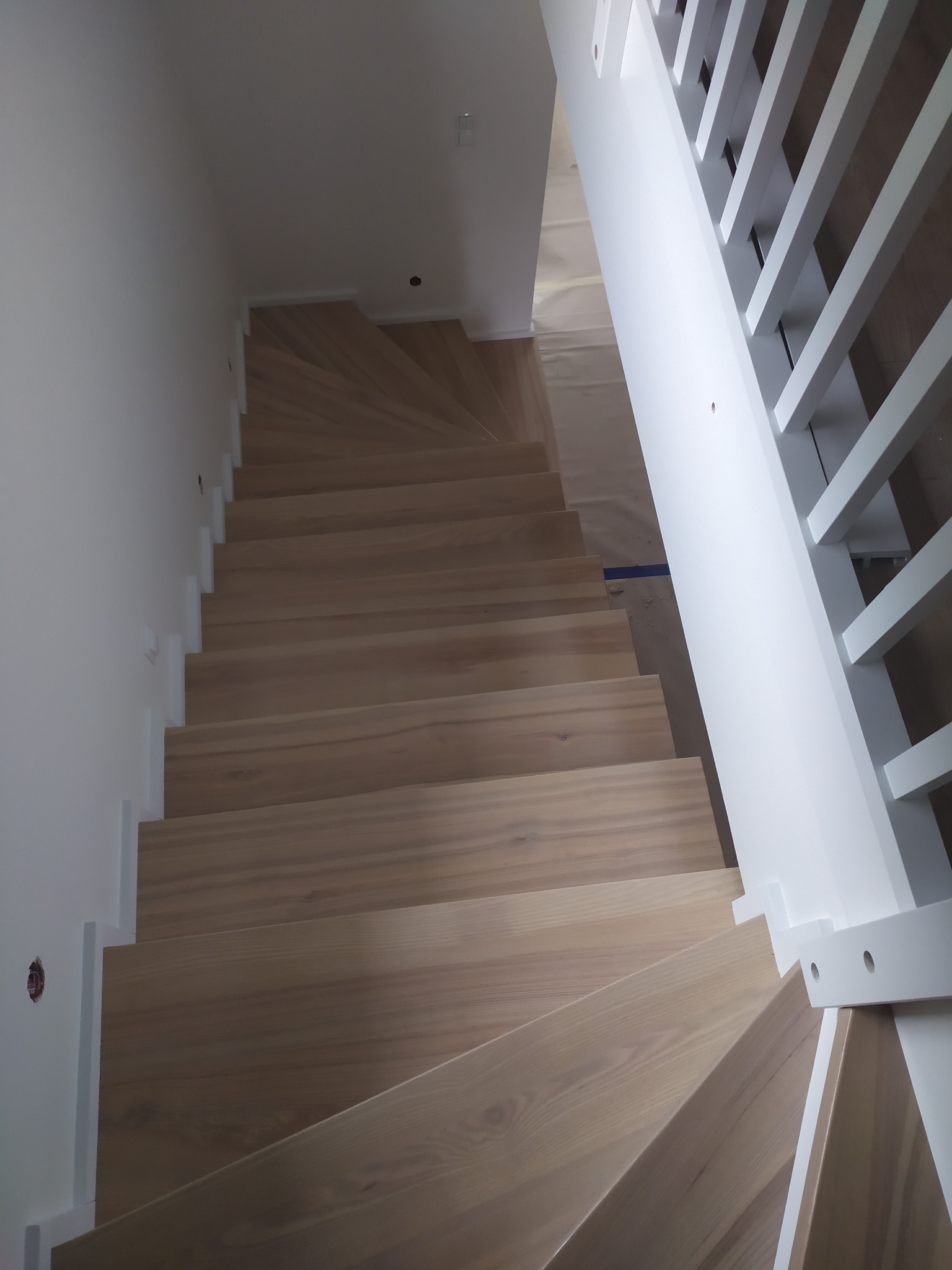 Jasne, drewniane schody z białymi balustradami i białymi listwami przypodłogowymi, widok z góry, częściowo zakryte folią ochronną.