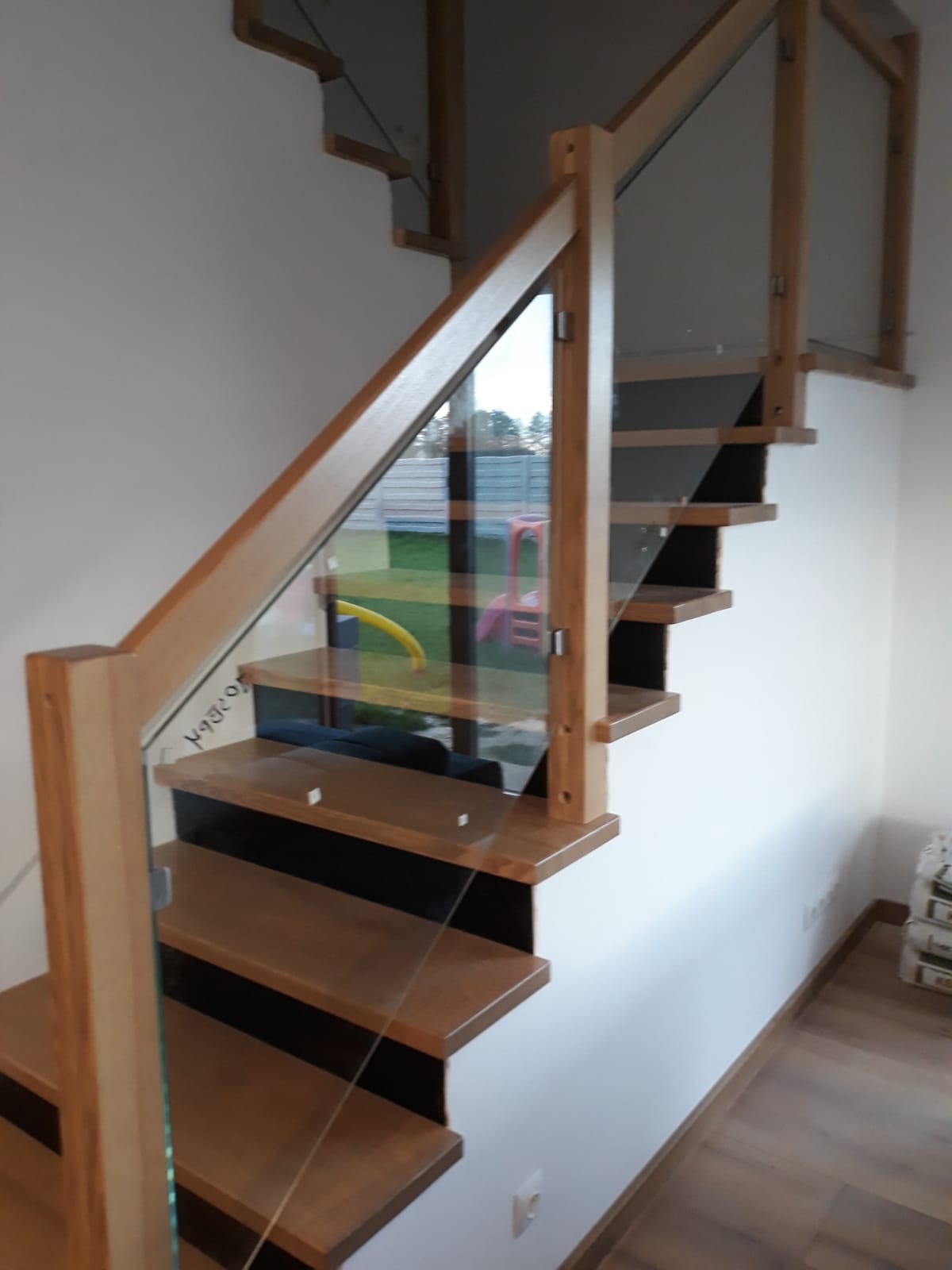 Nowoczesne schody z drewnianymi stopniami i szklaną balustradą, widoczne z boku, z elementami placu zabaw w tle za oknem.
