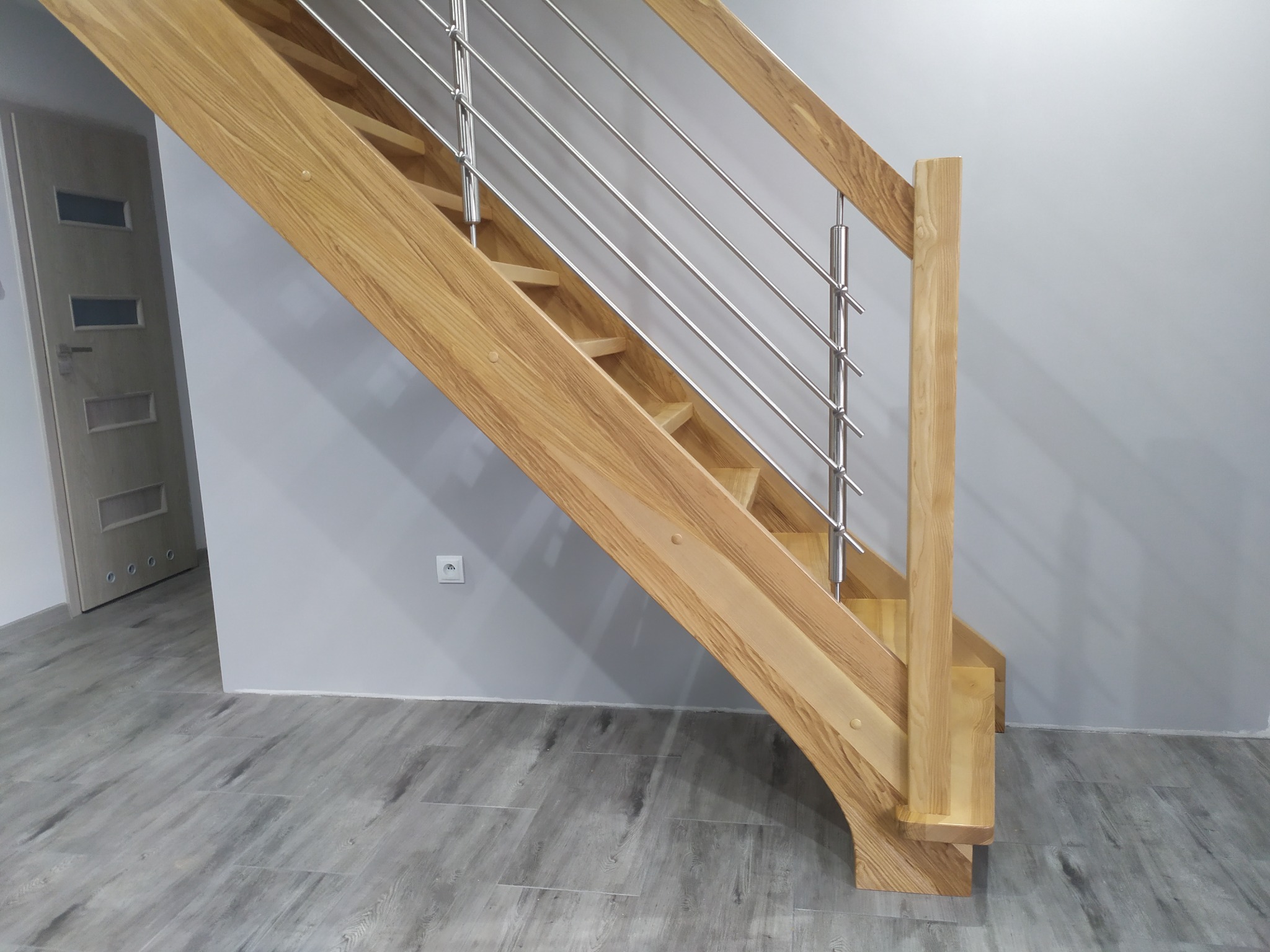 Jasne, dębowe schody z metalową balustradą, widoczne od dołu, z szarymi panelami podłogowymi i białą ścianą z gniazdkiem elektrycznym w tle.