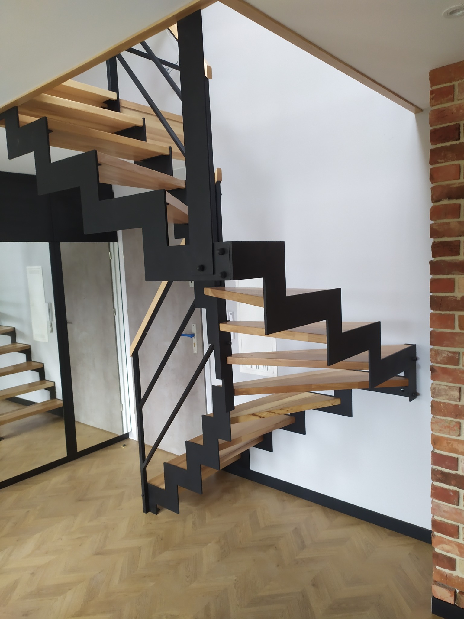 Nowoczesne schody metalowe z drewnianymi stopniami, czarne stalowe elementy konstrukcyjne, widok z dołu na parkiet w jodełkę, ściana z cegły w tle.