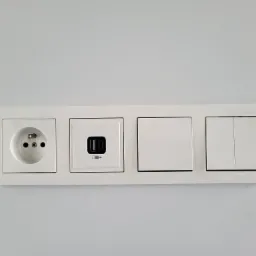 Biały panel z gniazdkiem elektrycznym, gniazdem USB z symbolem baterii i dwoma włącznikami światła na jasnej ścianie.