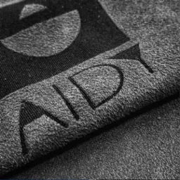 Czarno-białe zbliżenie na fragment szarej tkaniny z wytłoczonym, ciemnym napisem. Widoczna faktura materiału i precyzja wykonania detali liter.