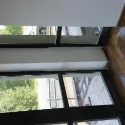 Świeżo pomalowana ściana nad oknem z czarną ramą, widok na zewnątrz. Podłoga wyłożona panelami w odcieniach brązu. Jasne, naturalne światło.