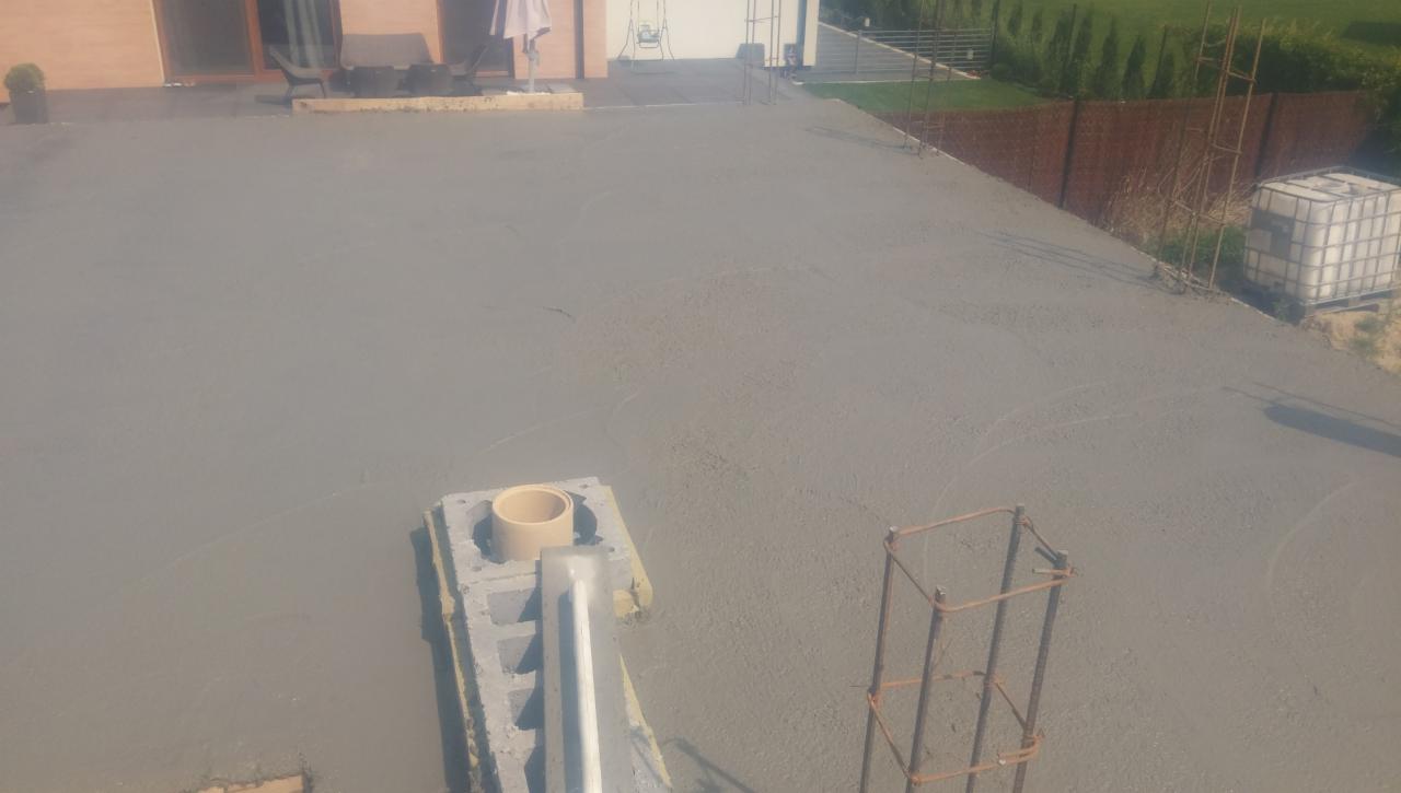 Świeżo wylany strop teriva, widoczne pustaki szalunkowe, rura wentylacyjna i zbrojenie, w tle dom w trakcie budowy i elementy ogrodu.