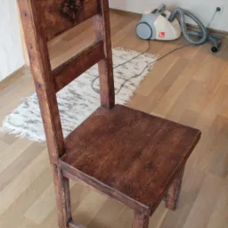 Rustykalne, drewniane krzesło z rzeźbionym ornamentem na oparciu, ustawione na podłodze z paneli. W tle odkurzacz i dywanik.