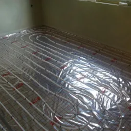 Białe rury ogrzewania podłogowego ułożone na folii z czerwoną siatką w jasnym pomieszczeniu, widoczne okno w górnym rogu.