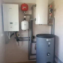 Zestaw urządzeń grzewczych w pomieszczeniu z beżowymi płytkami: pompa ciepła Panasonic, czerwony zbiornik wyrównawczy, biały zasobnik Galmet i szary zasobnik OEM Solar.