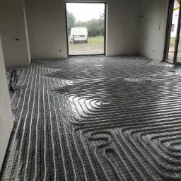 Układanie rur grzewczych na siatce w surowym wnętrzu, widoczny samochód dostawczy za oknem.